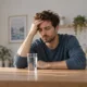 Un homme assis à une table pose une main sur son front, un verre d’eau devant lui.