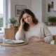 Une femme assise à une table pose une main sur sa tempe et l’autre sur son ventre.